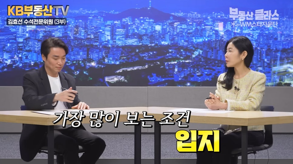 '스튜디오에서 두 명의 출연진이 대화를 나누는 장면'이다. 화면 하단에는 부동산을 선택할 때 사람들이 가장 많이 고려하는 핵심 조건으로 '입지'라는 단어가 노란색 글씨로 굵게 강조되어 나타나 있다.