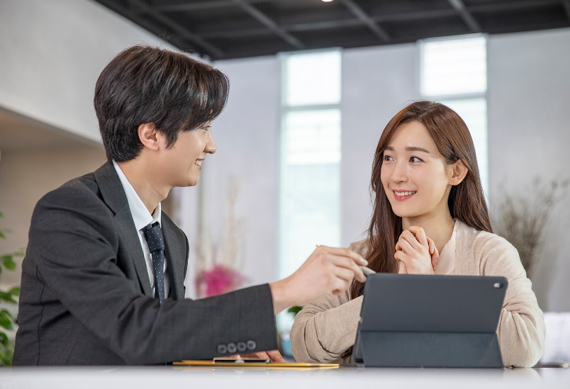 두 남녀가 취업 상담을 하는 듯함, 여자 앞에는 태블릿이 놓여 있으며, 남성이 테블릿 화면을 펜으로 가리키고 있음.