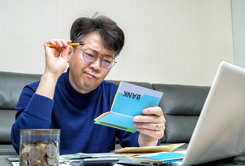 한 남성이 고민하는 듯한 모습으로 한 손에는 통장을 들고 있고, 다른 한 손에는 연필을 들고 머리에 대고 있으며, 책상 위에는 노트북이 열려 있음.