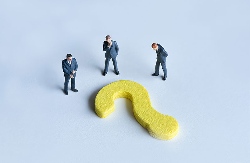 바닥에 놓여있는 노란색 물음표 근처에 정장차림을 한 세 남자가 서있다.