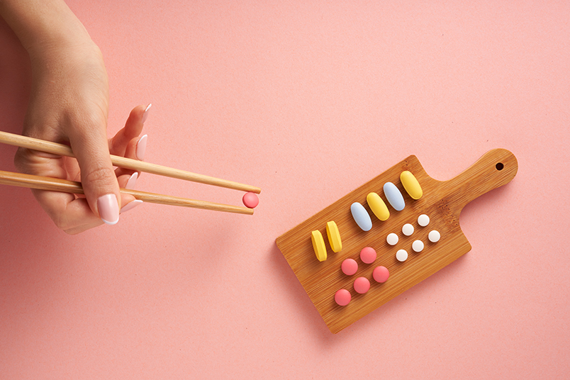 핑크색 바닥에 놓여진 도마, 나란히 모양별로 정렬된 형형색색 '알약' 들. 그중 동그란 핑크색 알약을 젓가락 끝으로 잡아 올리는 손.