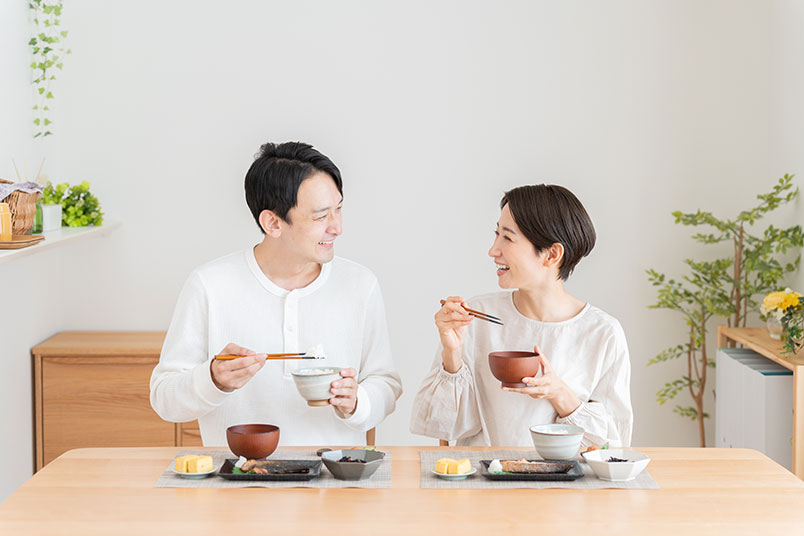 흰색 옷을 입은 부부가 식탁에서 마주보고 웃으며 식사를 하고 있다.