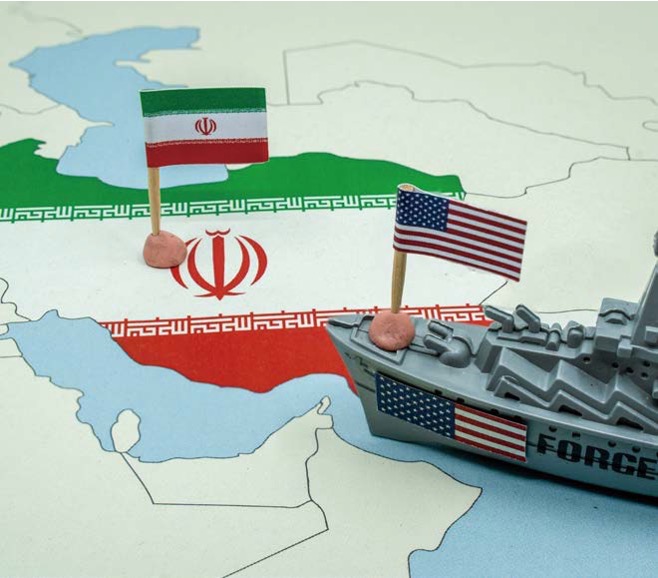 '중동 지도가 그려진 판 위에 이란 국기와 미국 국기가 꽂혀 있고, 성조기가 붙은 군함 모형이 이란 방향으로 배치되어 긴장감을 조성하는 모습'입니다.