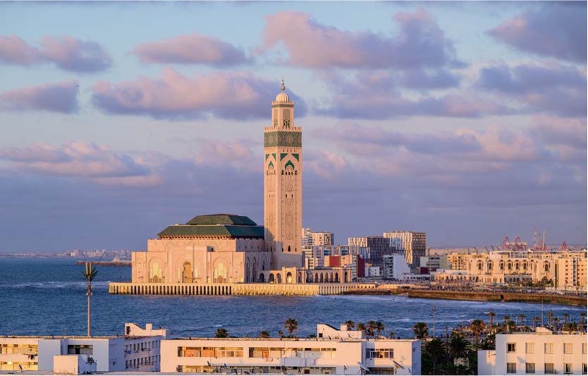 '카사블랑카 해안가에 우뚝 솟은 하산 2세 사원'의 전경이다. 세계에서 가장 높은 미나레트와 정교한 이슬람 건축 양식이 푸른 대서양 바다 및 도시 건물들과 대비를 이루는 평화로운 오후 모습이다.