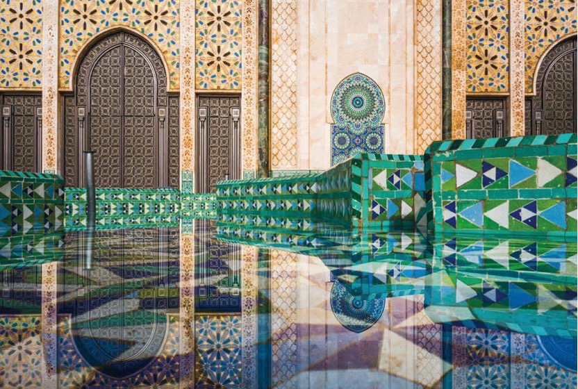 모로코 전통 문양인 젤리주(Zellij) 타일로 장식된 '사원 내부'이다. 기하학적인 패턴의 벽면과 에메랄드빛 타일이 맑은 물 위에 대칭으로 반사되어 화려하고 정교한 예술미를 보여주는 장면이다.