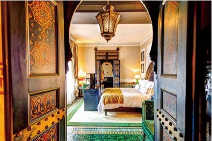 화려한 아라베스크 문양이 새겨진 나무 문 사이로 보이는 '라 마무니아 호텔의 고급스러운 객실이다. 천장의 정교한 랜턴 조명과 초록색 벨벳 소파, 전통 타일 바닥이 어우러져 마라케시 특유의 이국적이고 우아한 분위기를 자아낸다.