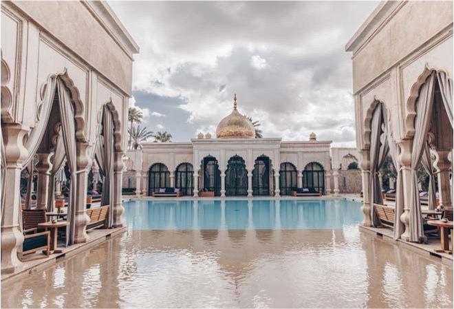 대칭을 이루는 흰색 아치형 기둥과 커튼이 드리워진 '테라스 사이로 펼쳐진 고급스러운 야외 수영장'이다. 중앙의 '황금빛 돔 지붕' 건축물과 맑은 수영장 물이 어우러진 마라케시의 '평화롭고 화려한 호텔 전경'이다.