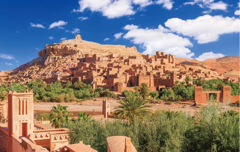 '붉은 진흙 벽돌로 지어진 고대 요새 도시'가 '거대한 바위산 기슭에 층층이 쌓인 모습'이 보인다. 푸른 하늘 아래 이국적인 야자수 숲과 흙빛 건축물들이 어우러진 '유네스코 세계문화유산 아이트벤하두'의 웅장한 풍경이다.