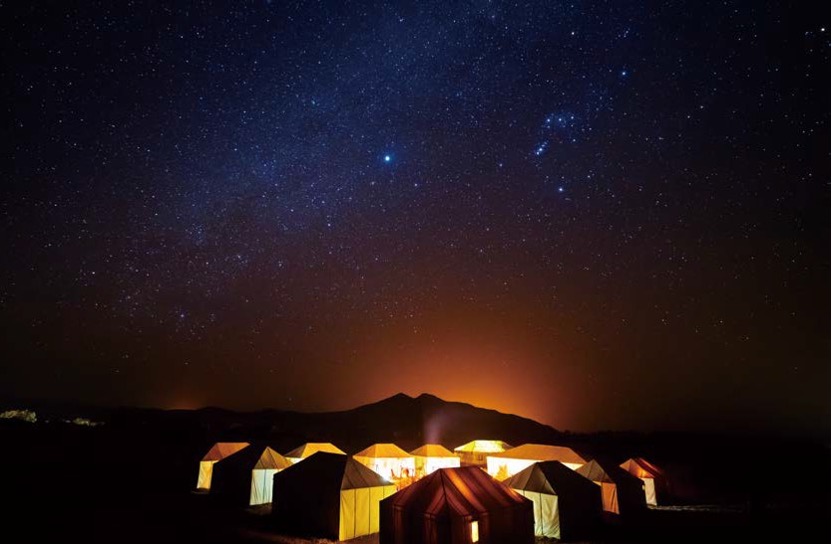 '쏟아질 듯한 별들이 가득한 밤하늘' 아래, '은은한 조명이 켜진 사막 텐트들'이 모여 있는 캠프 전경이다. 어둠 속 산의 실루엣과 텐트에서 새어 나오는 따뜻한 불빛이 어우러진 모로코 사하라 사막의 낭만적인 밤 풍경이다.