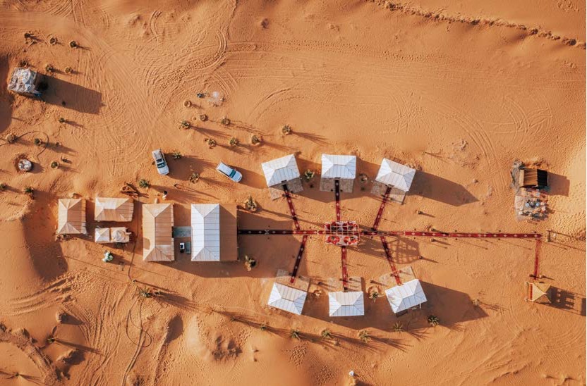 '붉은 사막 한가운데 기하학적으로 배치된 흰색 사막 텐트들'의 항공뷰이다. 중앙 광장을 중심으로 '방사형으로 뻗어 나간 카펫 길'과 주변의 지프차, 모래 위 발자국들이 어우러진 이색적이고 '체계적인 캠프 전경'이다.