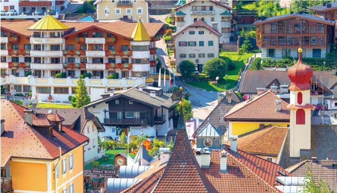 '돌로미티 마을의 아기자기한 건물들이 밀집'해 있으며, '붉은색 돔 형태의 종탑과 전통적인 알프스 양식의 지붕들'이 화려하고 따뜻한 마을 전경을 보여준다.