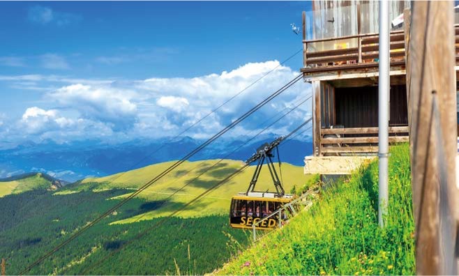 '노란색 세체다 케이블카가 푸른 숲과 광활한 산맥을 배경으로 가파른 경사를 오르내리며 돌로미티의 압도적인 고도감'을 생생하게 전달한다.