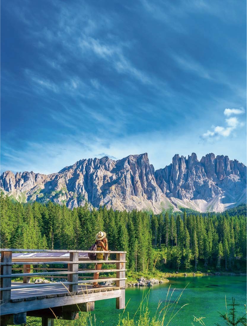'에메랄드빛으로 빛나는 카레자 호수 전망대에서 한 여행자가 모자를 쓰고 서서 거대한 라테마르 산맥의 암벽과 울창한 침엽수림을 감상'하고 있다.