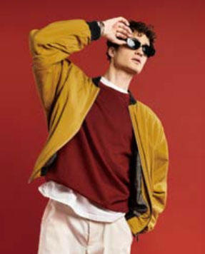 빨간색 니트와 겨자색의 아우터를 입은 남성 모델 한 명이 빨간색 배경 앞에서 자신감 있는 포즈를 취하고 있다.