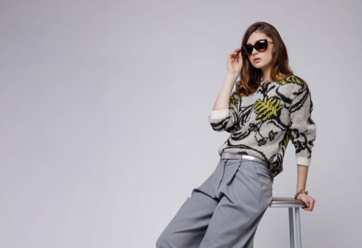 회색 배경에 선글라스를 쓰고 패턴이 큰 니트와 회색 바지를 입은 여성 모델이 의자에 앉아있다.