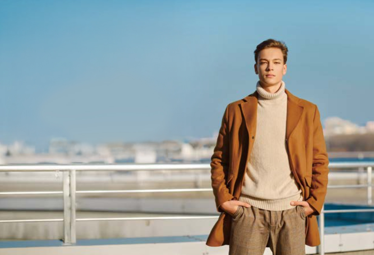 클래식한 울 코트, 베이지색 기본 니트와 슬랙스를 입은 남자 모델이 주머니에 손을 넣고 포즈를 취하고 있다.