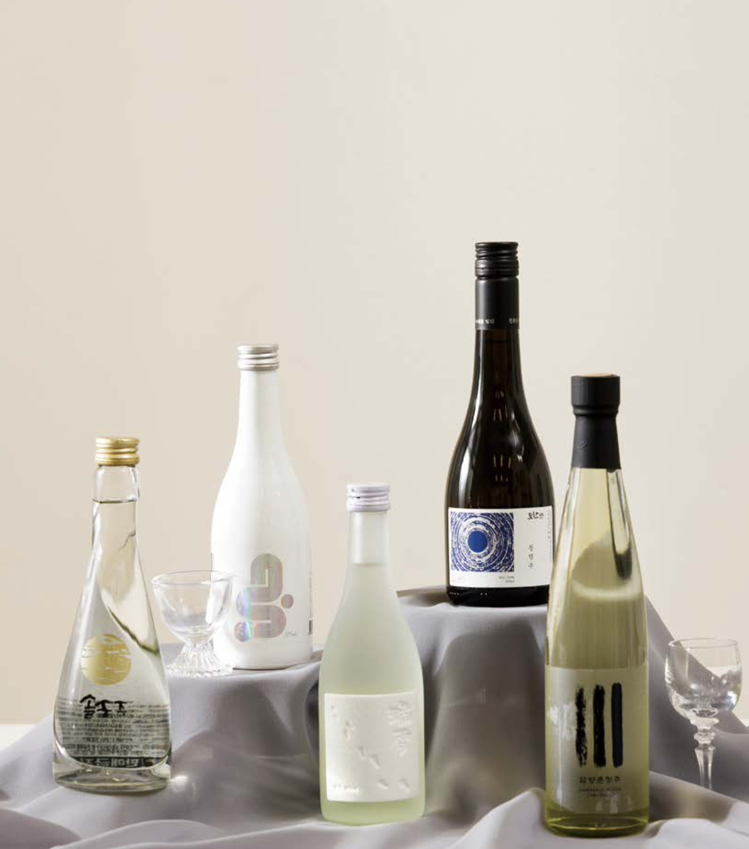 술마켓에서 추천하는 '맑은 술 5종'이 회색 천이 깔린 '받침대 위에 전시'되어 있으며, 주변에는 작은 술잔들이 함께 배치되어 있습니다.