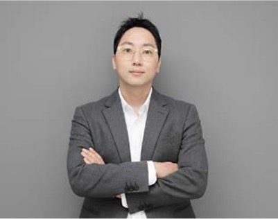 '회색 배경 앞에서 팔짱을 끼고 정면을 응시하는 안경 쓴 남성'의 비즈니스 프로필 사진이다.