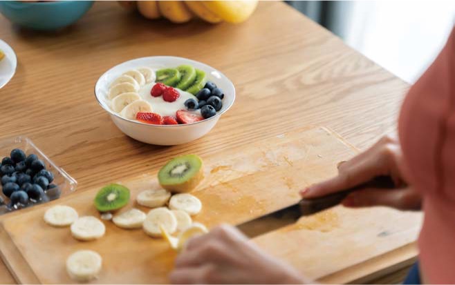 나무 테이블 위에 비슷한 톤의 나무 도마에서 '바나나와 키위를 썰어 요거트 볼에 과일 토핑을 담는 손길'이다.