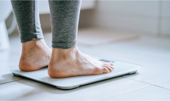 '회색 레깅스를 입은 사람'이 '흰색 디지털 체중계 위에 맨발로 올라가 무게를 재는 모습'이다.
