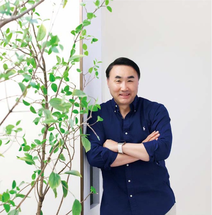 '남색 셔츠를 입은 남성이 나무 옆에 기대어 팔짱을 낀 채 인자한 미소를 지으며 정면을 응시'하고 있다. 배경은 깔끔한 흰색 벽이다.