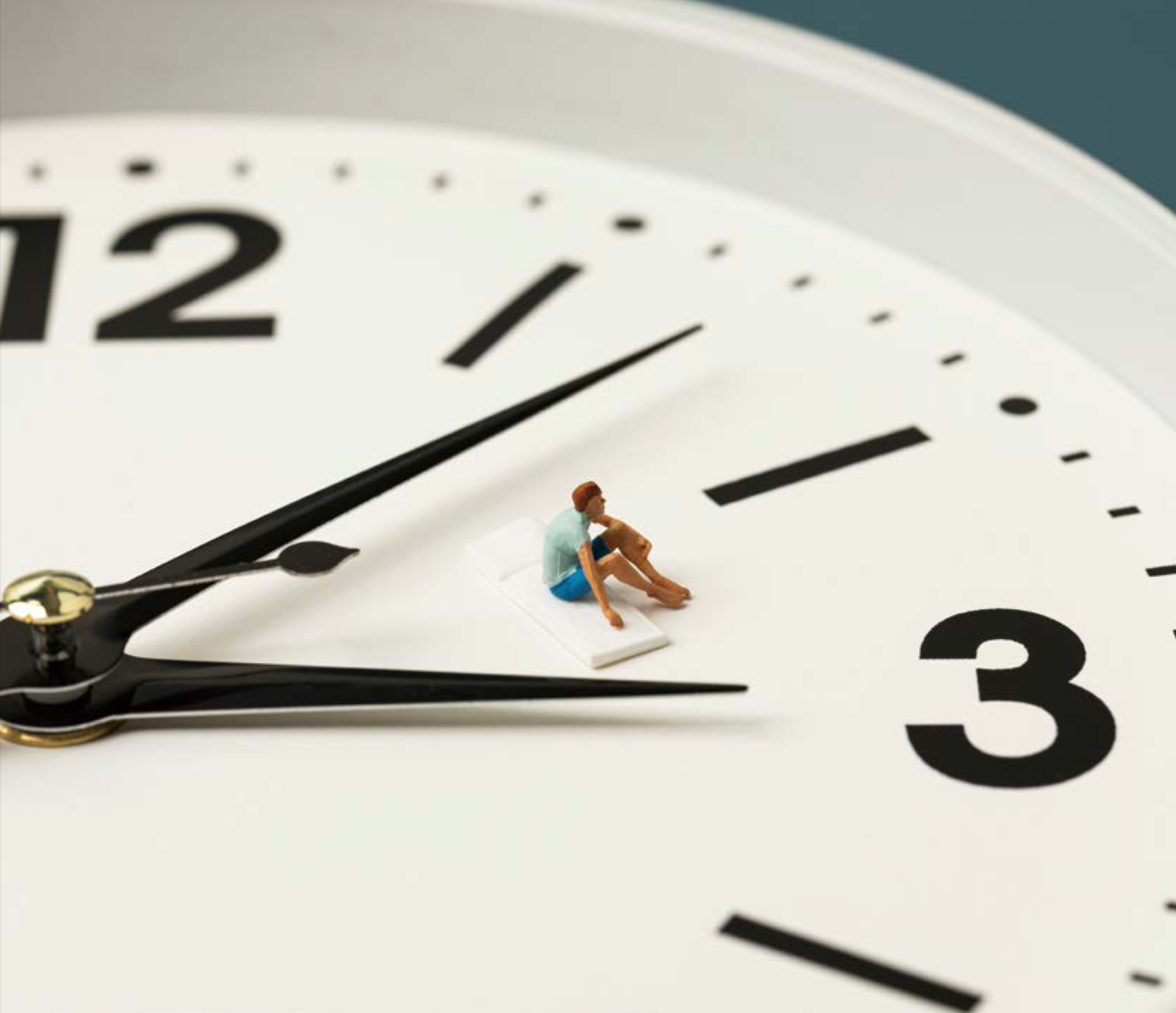 커다란 아날로그 '시계 판위에 아주 작은 사람 모형'이 '앉아 휴식을 취하고 있는 미니어처 연출 사진'입니다. 시곗바늘과 숫자판을 배경으로 시간의 흐름을 시각적으로 표현했습니다.