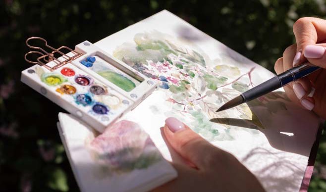 '휴대용 미니 팔레트와 붓을 사용하여 화사한 꽃과 잎사귀를 수채화로 그려내고 있습니다.' 분홍색과 초록색 물감이 종이 위에서 부드럽게 번지며 생동감 넘치는 꽃의 형태를 완성해 갑니다.