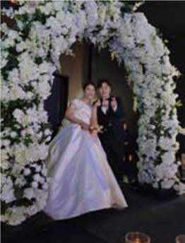 '꽃 아치 아래의 신랑 신부가 서로를 마주 보며 웃고 있고 주변에 분홍 꽃잎'이 흩날린다. 영상 편집 중 초반 이미지이다.