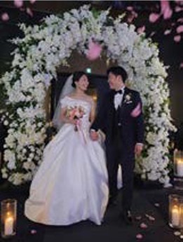 '꽃 아치 아래의 신랑 신부가 서로를 마주 보며 웃고 있고 주변에 분홍 꽃잎'이 흩날린다. 영상 편집 중 중반 이미지이다.