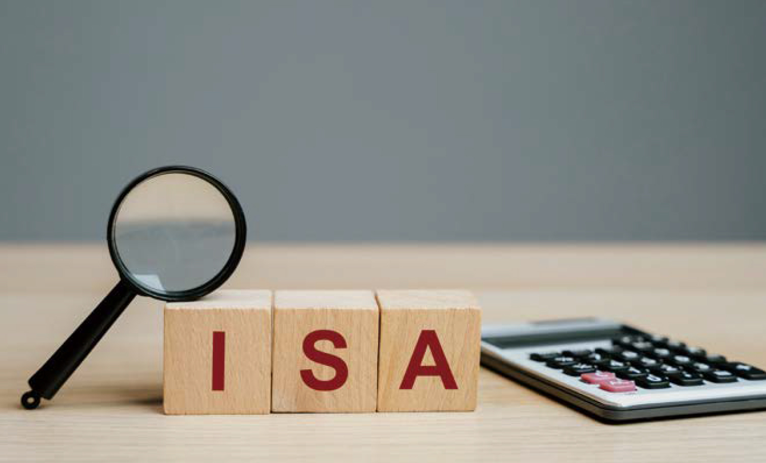 원목 책상 위에 올려져 있는 나무 블록에 ISA가 적혀있고 그 옆에는 돋보기와 계산기가 놓여있다.