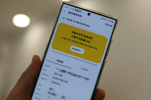 왼손으로 쥔 스마트폰, 'KB차차차 앱' 의 '팔아줘차차차' 페이지를 보여주고 있음. '내차고 서비스'' 등록 차량의 '사진 등록' 단계를 보여주고 있음.