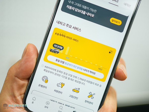 왼손에 스마트폰을 든 상태, ;KB차차차' 앱 내 '신차와 중고차' 사이의 '감가' 정도를 확인할 수 있는 서비스를 보여주고 있음.