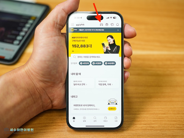 나무 책상 위, 왼손으로 스마트 폰을 잡고 'KB차차차' 앱의 '내차고 등록' 을 안내하는 이미지.