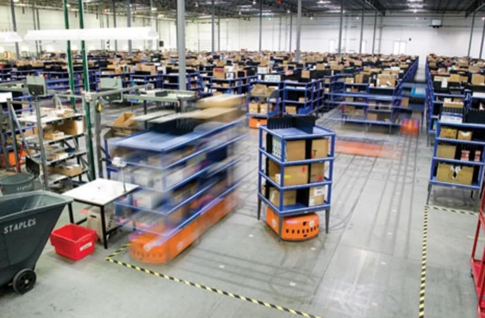 아마존 풀필먼트 센터 내에서 오렌지색 물류 로봇 '키바(Kiva)'들이 선반을 운반하며 분주하게 움직이는 모습입니다. 넓은 창고 부지에 수많은 상품 선반이 정렬되어 있고, 로봇을 통한 자동화된 물류 시스템의 현장을 보여줍니다.