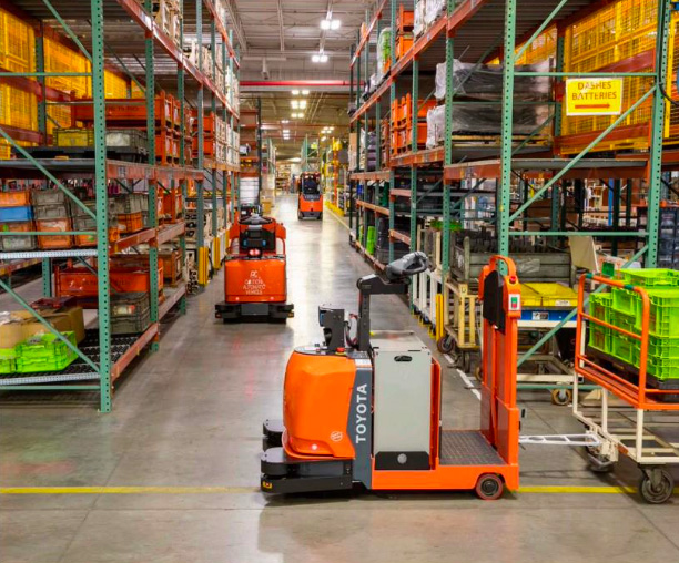 물류 창고 내 선반 사이로 '토요타(TOYOTA)의 오렌지색 자동 유도 차량(AGV)들'이 '이동하는 모습'입니다. 산업 현장에서 자재를 운반하며 자동화 시스템을 구현하는 장면을 담고 있습니다.