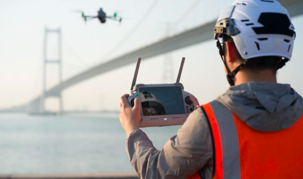 '안전 조끼와 헬멧을 착용한 작업자'가 대형 교량 근처에서 컨트롤러를 이용해 '드론을 원격 조종하는 모습'입니다. 시설물 점검을 위해 드론을 운용하는 현장을 보여줍니다.