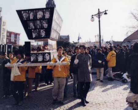 '스크린쿼터 축소에 반대하는 사람들이 거리에서 행진하는 사진'이다. 참가자들이 인물 사진을 붙인 대형 구조물과 가로로 긴 막대를 들고 이동하며, 뒤쪽까지 많은 시위 인파가 이어진다.