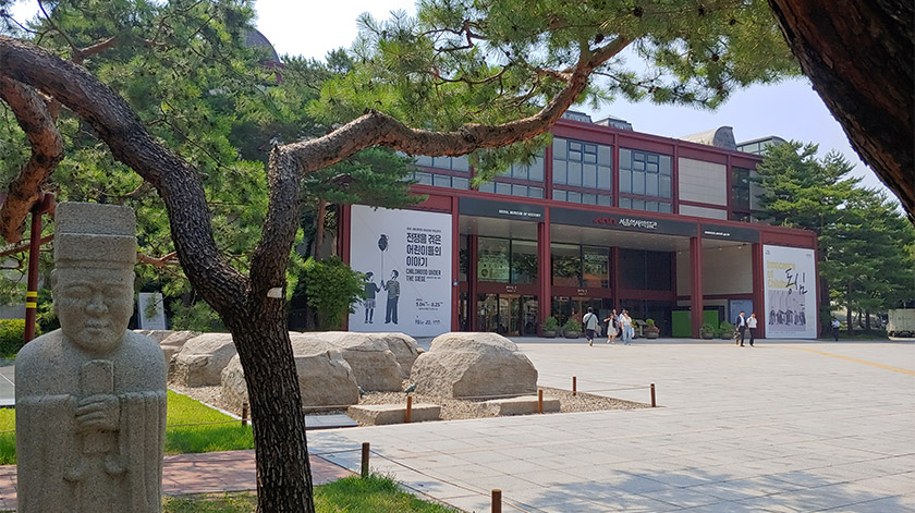 박물관 전시실의 미니멀한 화이트 테이블 위에 놓인 다양한 크기와 모양의 한국 전통 청화백자들. 배경에는 2027년 유료 전환 안내와 박캉스 코스 추천 텍스트가 포함됨