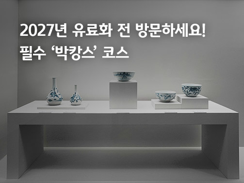 박물관 전시실의 미니멀한 화이트 테이블 위에 놓인 다양한 크기와 모양의 한국 전통 청화백자들. 배경에는 2027년 유료 전환 안내와 박캉스 코스 추천 텍스트가 포함됨
