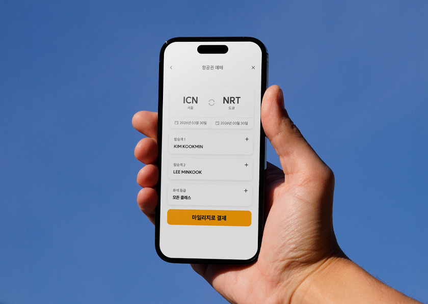스마트폰을 든 손. 스마트폰 화면에는 항공권 예약 화면이 있고, 마일리지로 결제라고 쓰인 버튼이 하단에 보인다. 