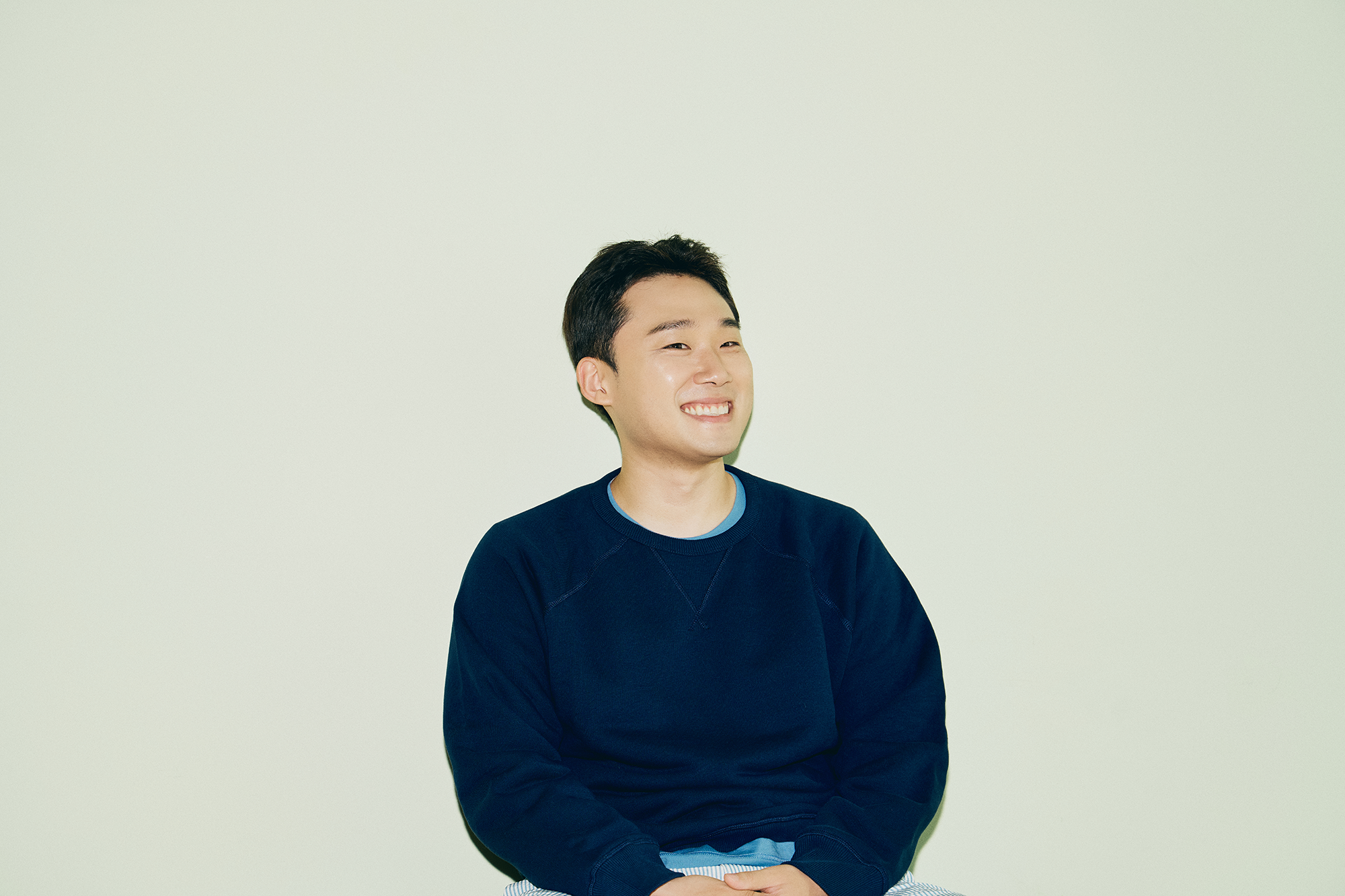 네이비색 상의를 입고 흰 벽을 배경으로 앉아 미소 짓고 있는 자취남