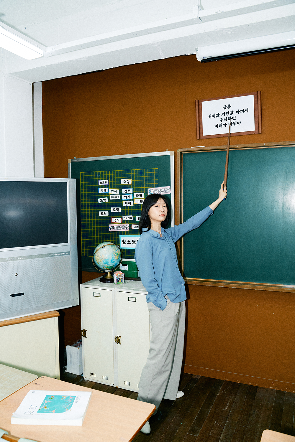 옛 초등학교 교실을 배경으로 칠판 앞에 서서 급훈을 가리키고 있는 슈카월드PD 니니. 급훈에는 '커피값 치킨값 아껴서 주식하면 미래가 바뀐다'라고 쓰여 있다.