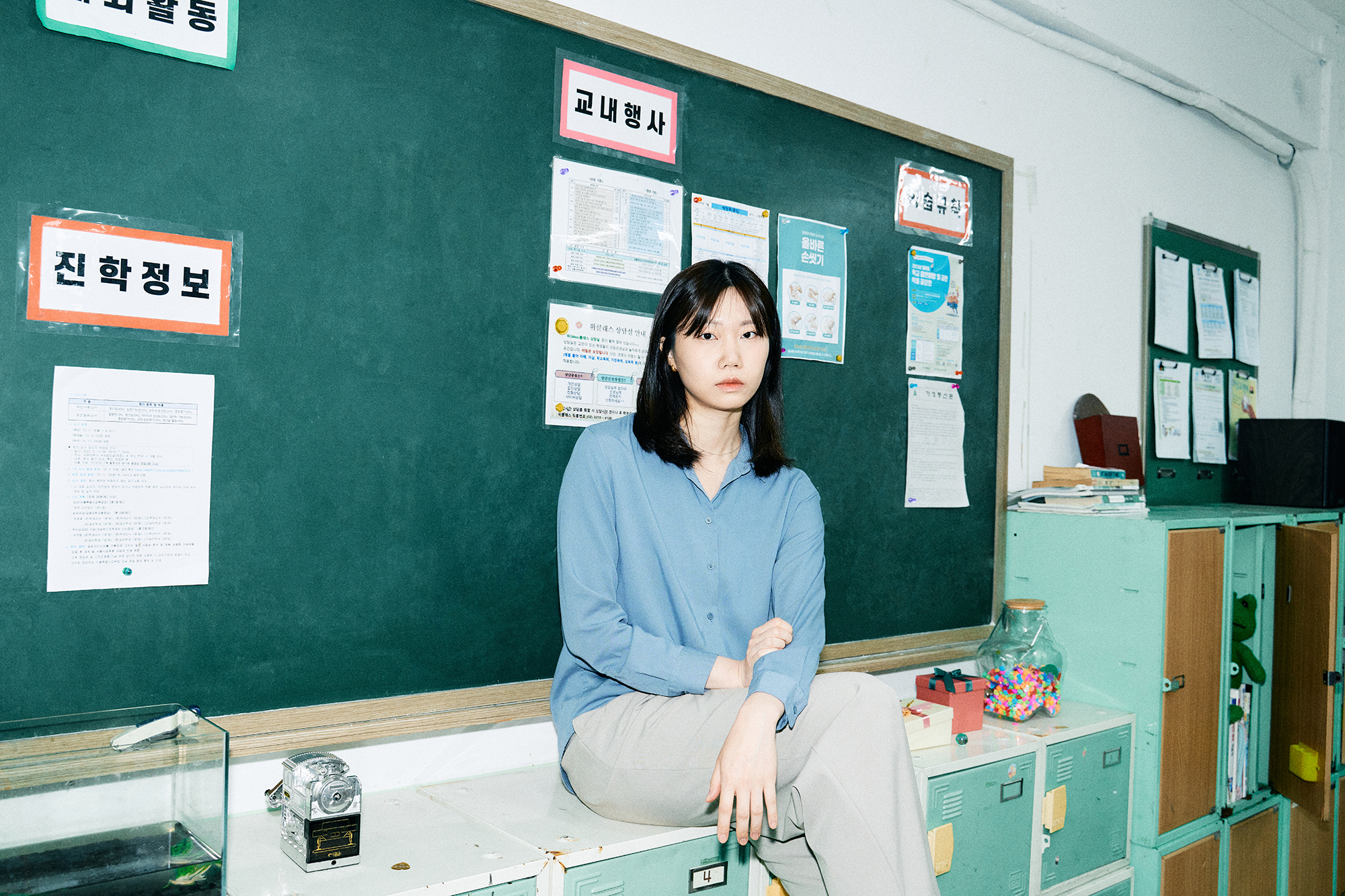 옛 초등학교 교실을 배경으로 사물함에 걸터앉아 정면을 바라보고 있는 슈카월드PD 니니