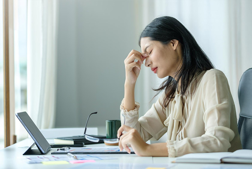 책상 앞에 앉아 미간을 짚으며 피곤해하는 여성의 모습이다.
